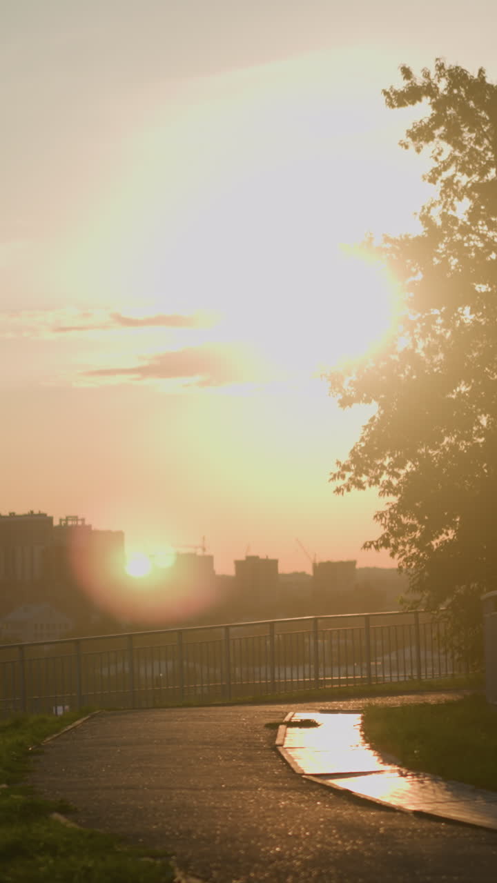vista del paisaje urbano al atardecer con personas caminando por el camino cerca de la barandilla de hierro, grandes árboles proyectando sombras, la cálida luz del sol iluminando los edificios urbanos en el fondo