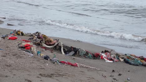 Montones-De-Ropa-Y-Telas-Desechadas,-Moda-Rápida,-Arrastradas-A-La-Playa-De-Arena-Desde-El-Océano-Con-Suaves-Olas-En-Un-Destino-De-Isla-Tropical