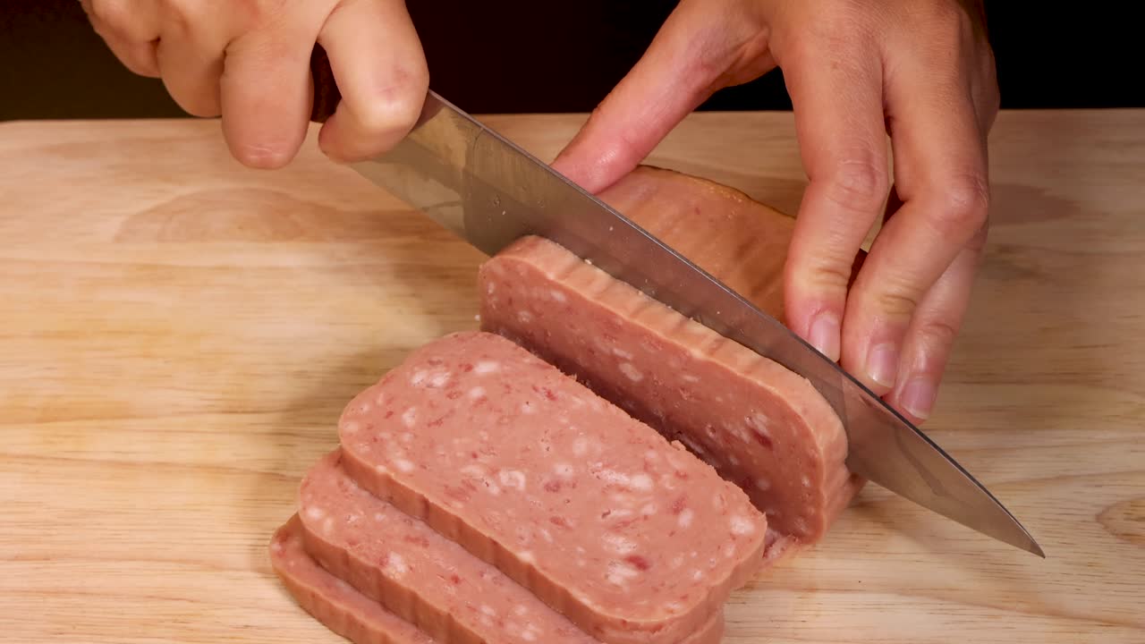 Hands use a large kitchen knife to slice a block of canned processed meat into even pieces on a wooden cutting board under warm lighting