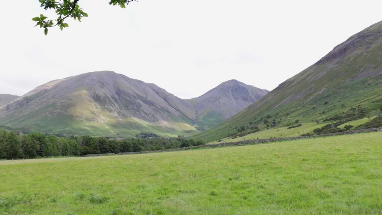 Southern Fells, Cumbria, panoramica verso destra attraverso un ampio prato verde verso scoscesi pendii montuosi e una linea di alberi, catturando l'atmosfera della campagna aperta sotto una soffice copertura nuvolosa.