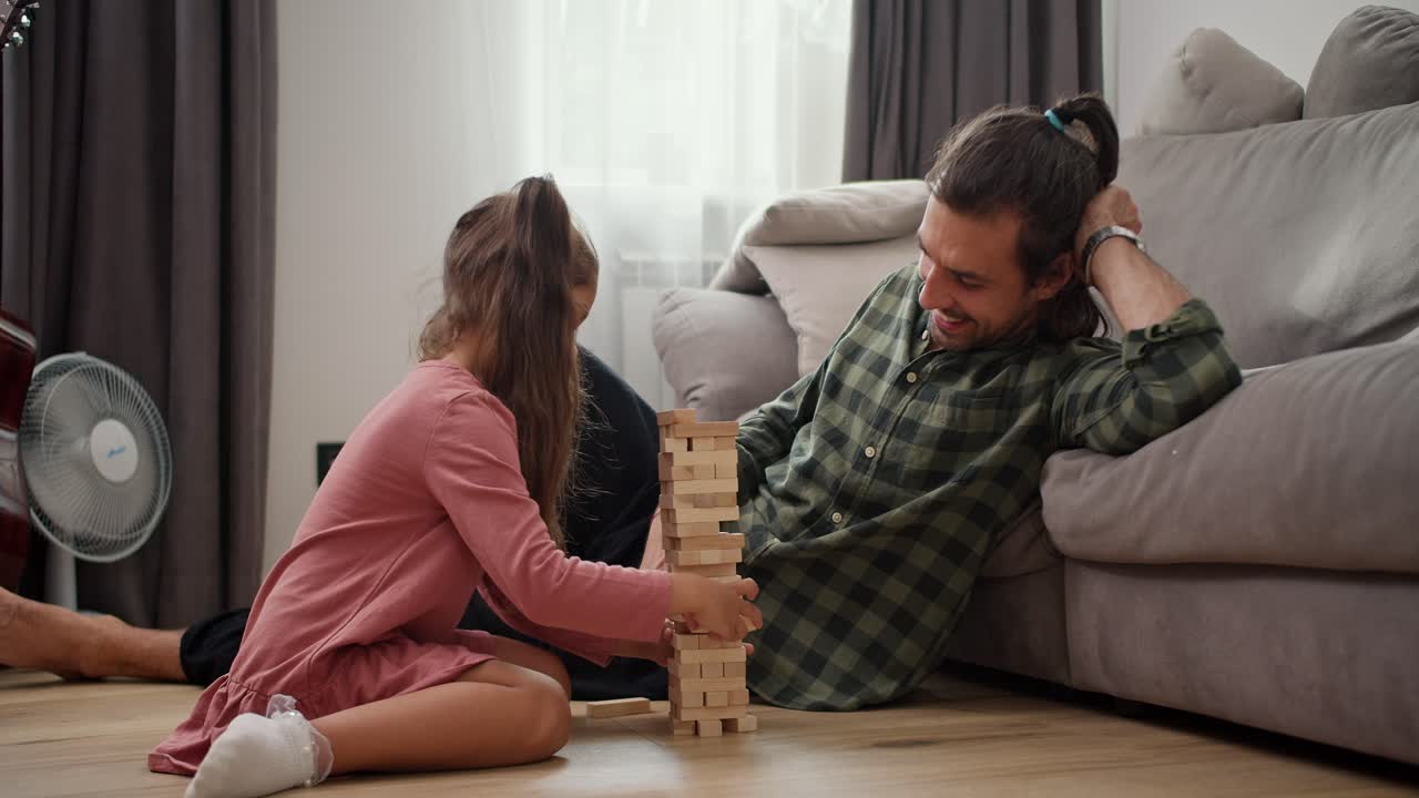 Side view of a little brunette girl in a pink dress plays with her brunette father in a checkered green shirt the board game Jenga and when she pulls out one of the bricks the Jenga tower falls