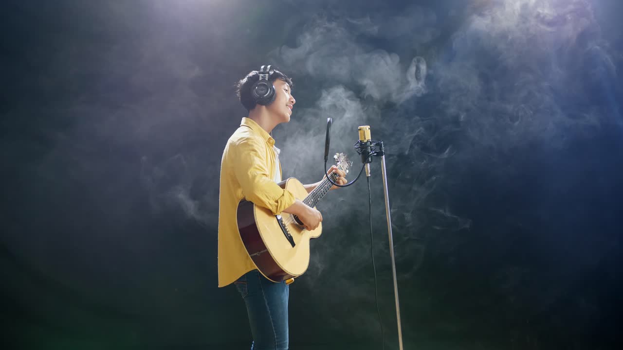하 연기 검은 배경에 기타를 연주하고 콘덴서 마이크로 노래하는 헤드폰을 가진 젊은 아시아 소년의 측면 시각