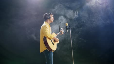 vista lateral de un joven asiático con auriculares tocando una guitarra y cantando en un micrófono condensador en el fondo blanco de humo negro
