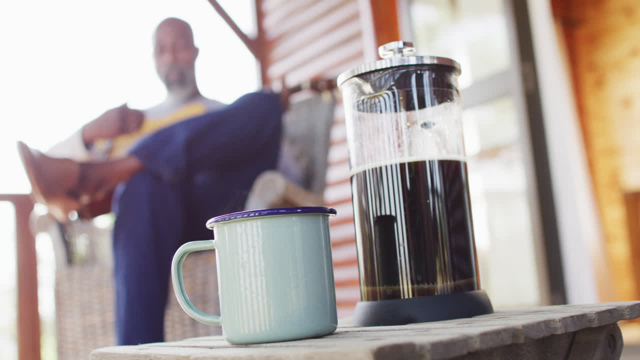 Mug of coffee over senior african american man in log cabin, playing guitar on balcony, slow motion