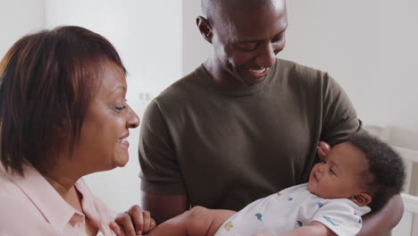 Proud-Grandmother-With-Adult-Son-Cuddling-Baby-Grandson-In-Nursery-At-Home