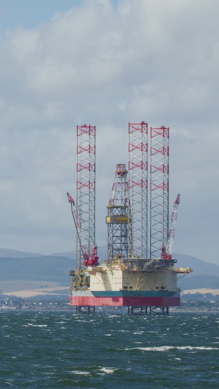 A stationary offshore oil drilling rig stands in choppy, wind-swept waters near Cromarty, Scotland, under bright daylight with distant hills visible