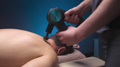 masajeador de percusión de primer plano en las manos de un masajista hace un masaje de los hombros y la parte superior de la espalda a un cliente masculino acostado