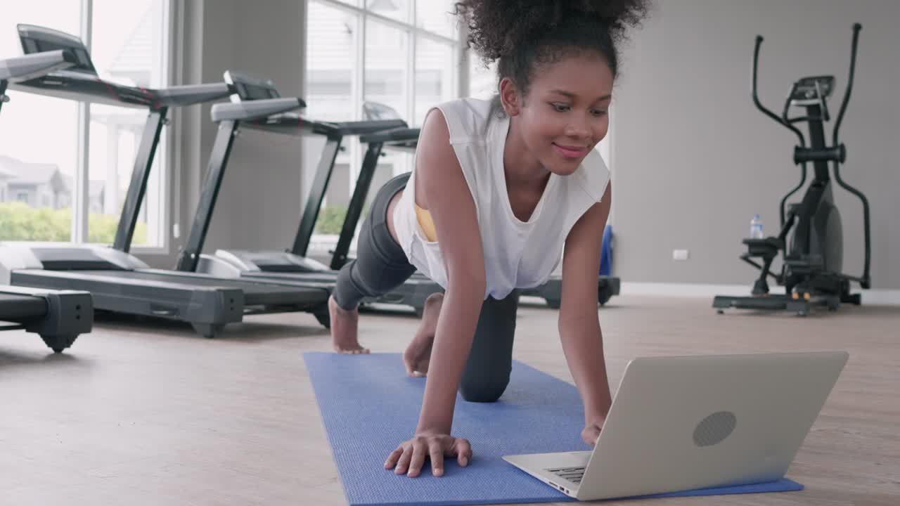 joven negra atractiva y en forma haciendo ejercicio de yoga en el gimnasio. mujer afroamericana en la lección de portátil practicando la postura de tabla de yaga en la clase de internet en línea social. concepto de estilo de vida saludable
