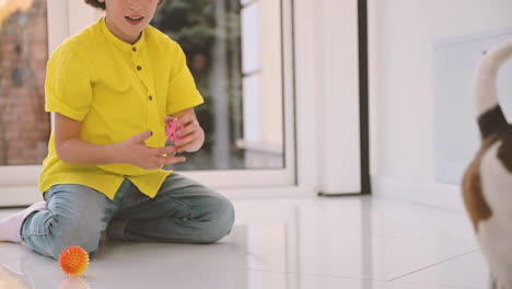 Blond-Boy-With-Curly-Hair-Kneeling-On-The-Floor-While-Playing-With-Balls-With-His-Dog