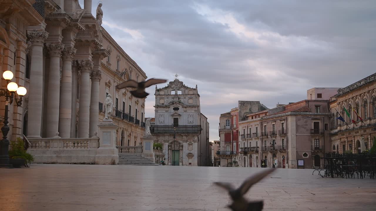 Piccioni volano sopra la piazza principale vuota, Piazza Duomo nell'isola di Ortigia, all'alba nella provincia di Siracusa in Sicilia.