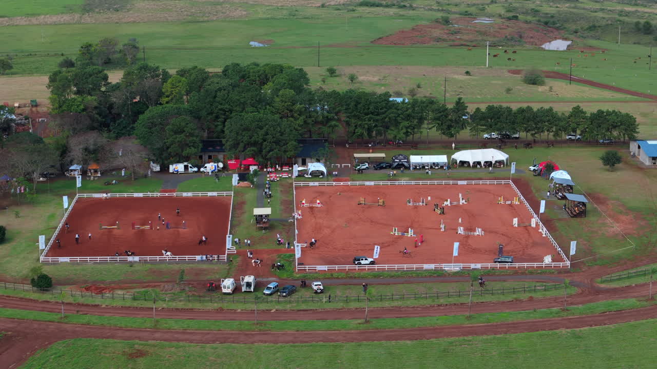 Smooth right-sliding drone shot captures riders training in vibrant red soil arenas at an equestrian center, showcasing movement against earthy tones. Ideal for sports promotions.