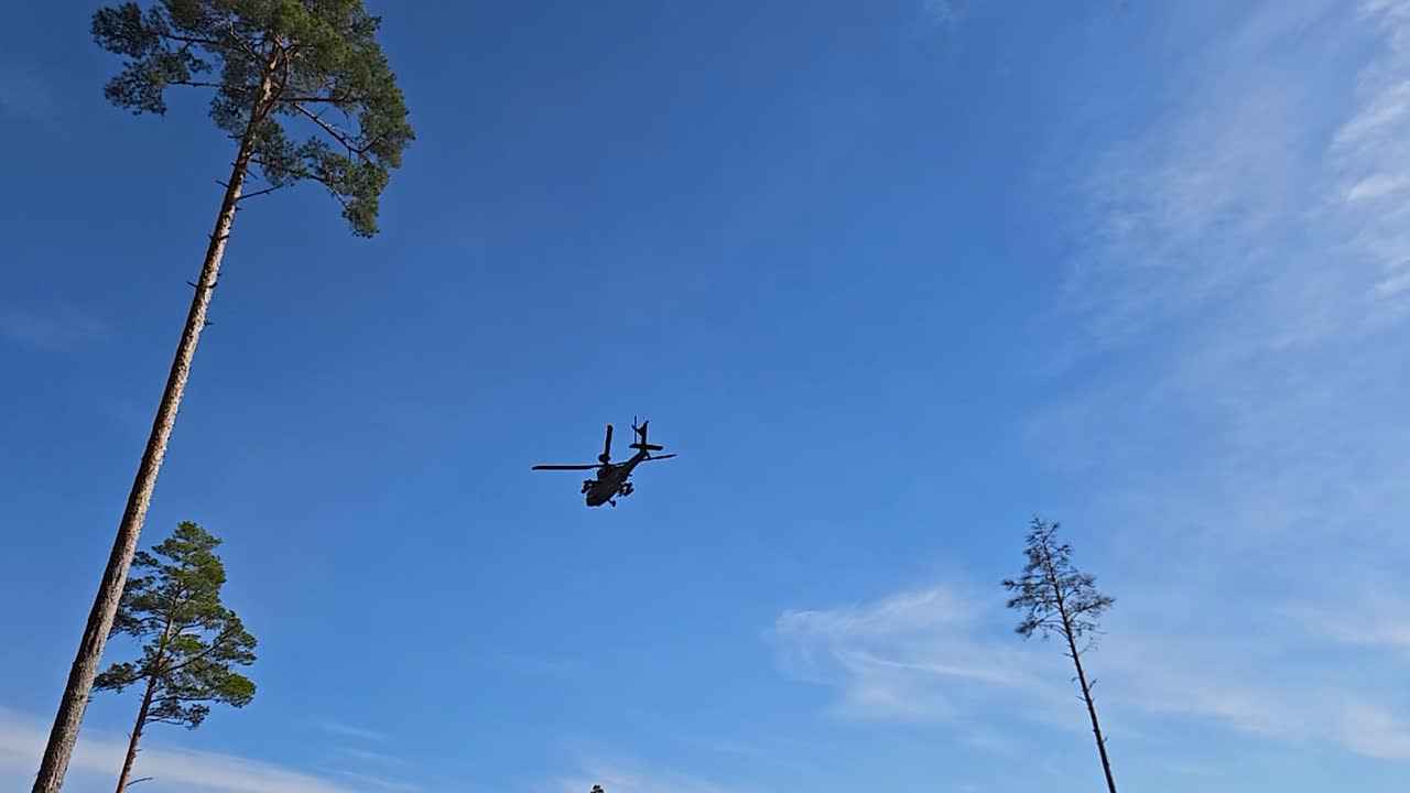 Rare military footage of a Boeing AH-64 Apache helicopter flying over a sandy and green grassy infanty trenches at a war or battle zone during a sunny day. Tall pine trees and treeline visible in back