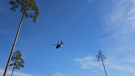 rare military footage of a boeing ah-64 apache helicopter flying over a sandy and green grassy infanty trenches at a war or battle zone during a sunny day. tall pine trees and treeline visible in back