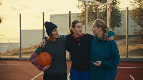 a trio of happy girls basketball players with an orange ball in a sports uniform stand on a street red court in the morning and communicate hugging