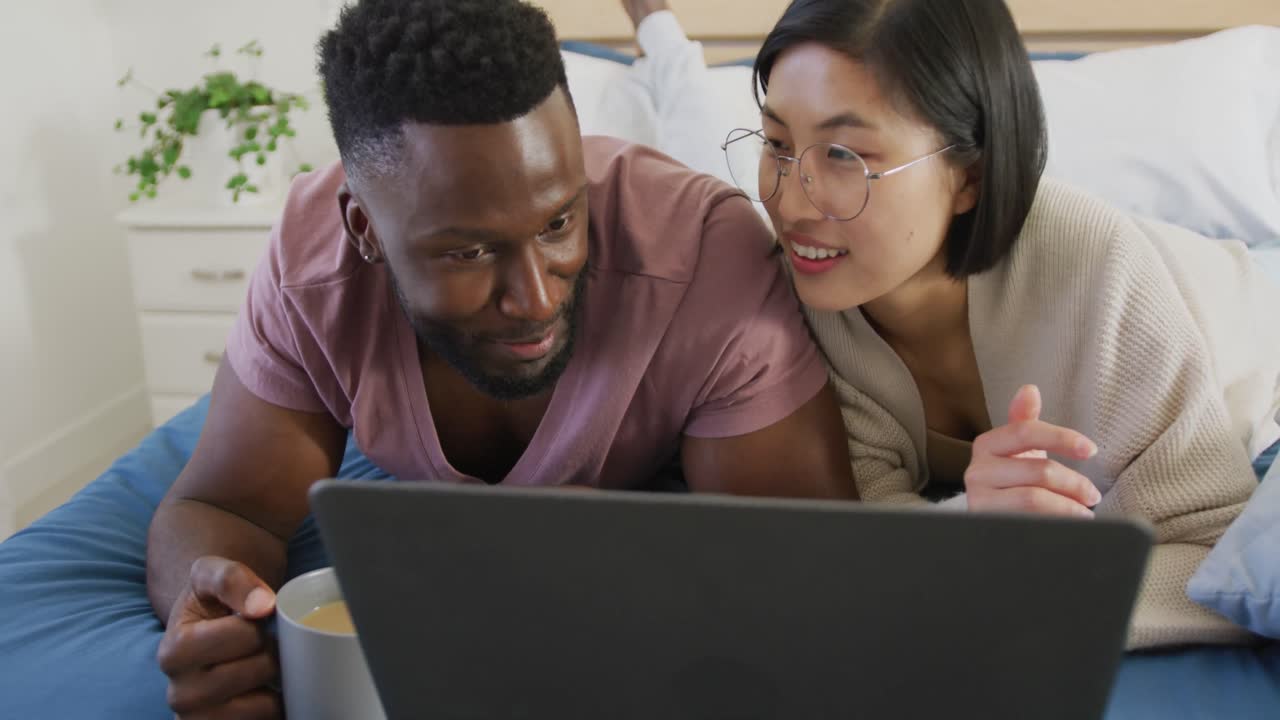 Happy diverse couple using laptop and lying in bedroom