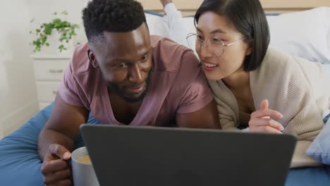 happy diverse couple using laptop and lying in bedroom