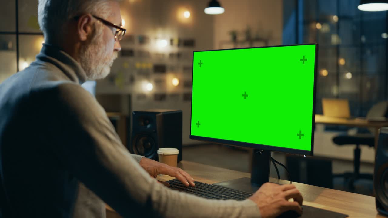 diseñador creativo de mediana edad sentado en su escritorio utiliza computadora de escritorio con pantalla de maqueta verde. por la noche dos empleados creativos trabajando en computadoras en la oficina con vista de ventana del paisaje urbano