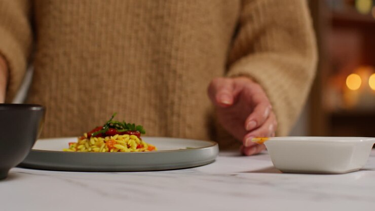 Primo piano di donna a casa in cucina che prepara un pasto sano vegetariano o vegano, cospargendo erbe aromatiche su pasta orzo e pomodori arrostiti