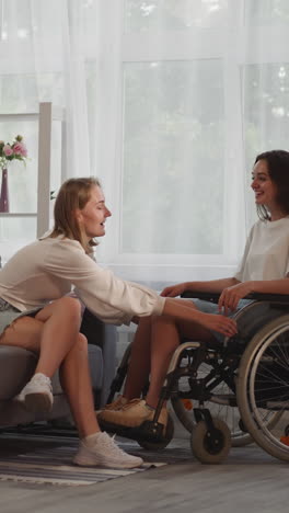 caring woman moves wheelchair with girlfriend with disability close sitting on sofa near window. happy lesbian ladies enjoy talking at home