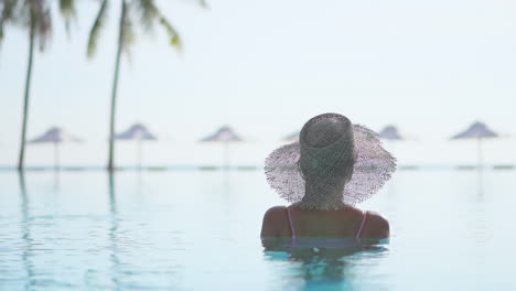 a woman with her back to the camera in chest-high pool water looks out toward the beach umbrellas and ocean