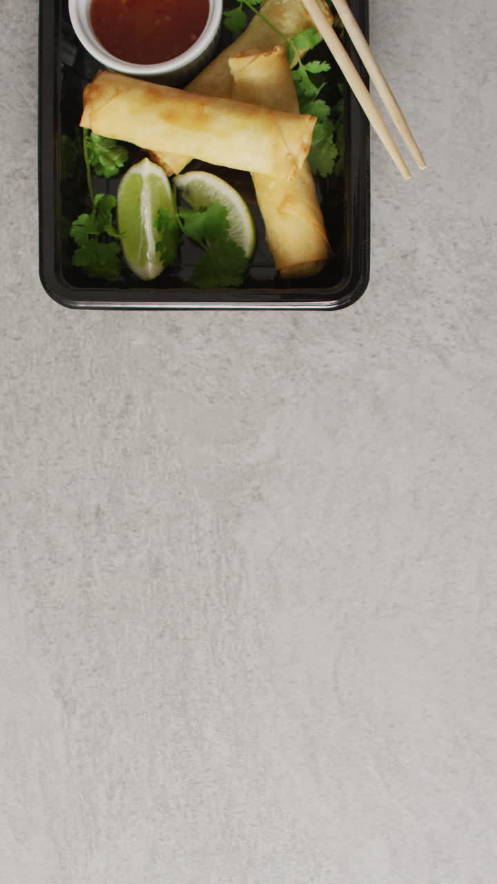 video vertical de un plato con rollos de primavera y salsa de chile con palillos sobre un fondo gris