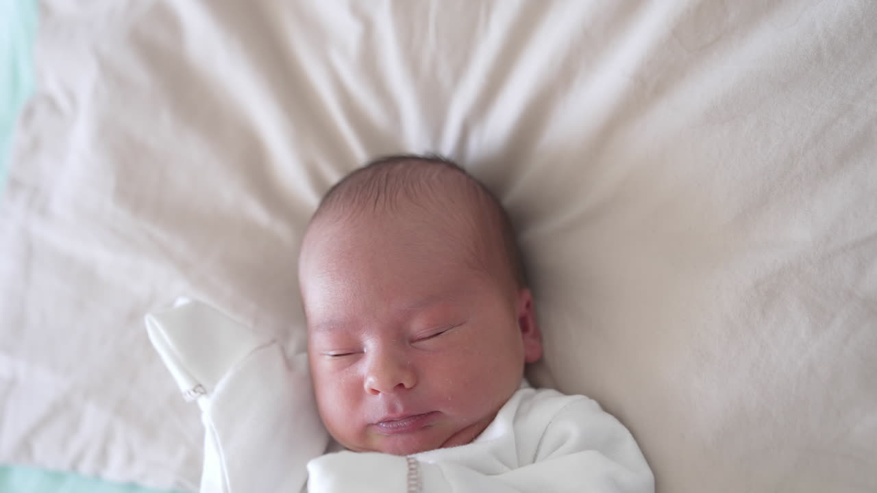 Infant baby. Portrait of a newborn baby in white clothes lying the bed. Cute baby boy waking up and blinking his eyes. Close-up.
