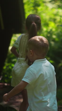 cute little girl and boy look around near arbor in shady woodland park slow motion. healthy brother and sister in summer forest. active weekend