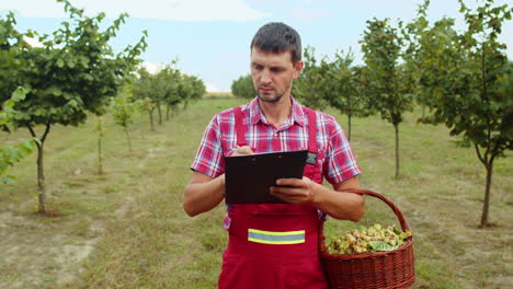 Gardener-agronomist-man-farmer-analyzing-hazelnuts-tree-rows-in-garden-making-notes-in-journal