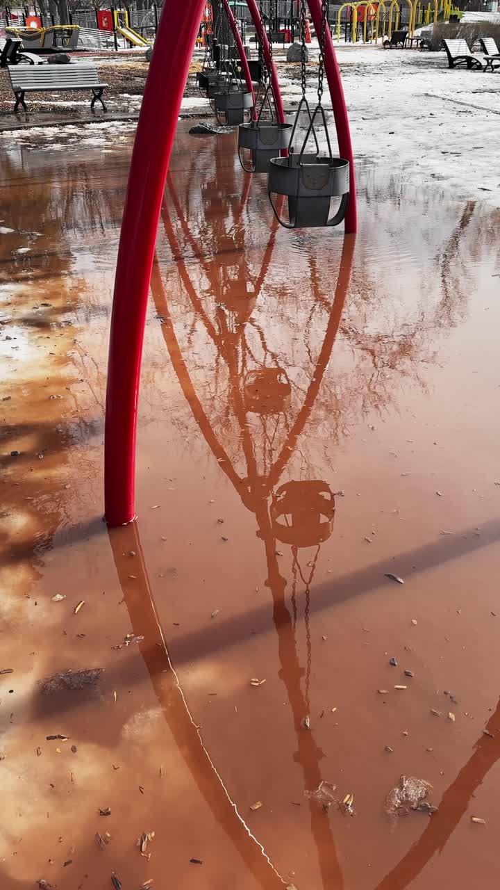 Lo scioglimento primaverile della neve trasforma le altalene vuote di un parco giochi in un parco cittadino in una scena di alluvione riflettente, lasciando la superficie di gioco invasa da acqua stagnante mentre l'inverno cede il passo alla primavera.
