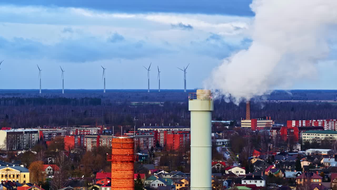Städtisches Industriegebiet mit qualmenden Schornsteinen und Windrädern am Horizont unter einem bewölkten Himmel