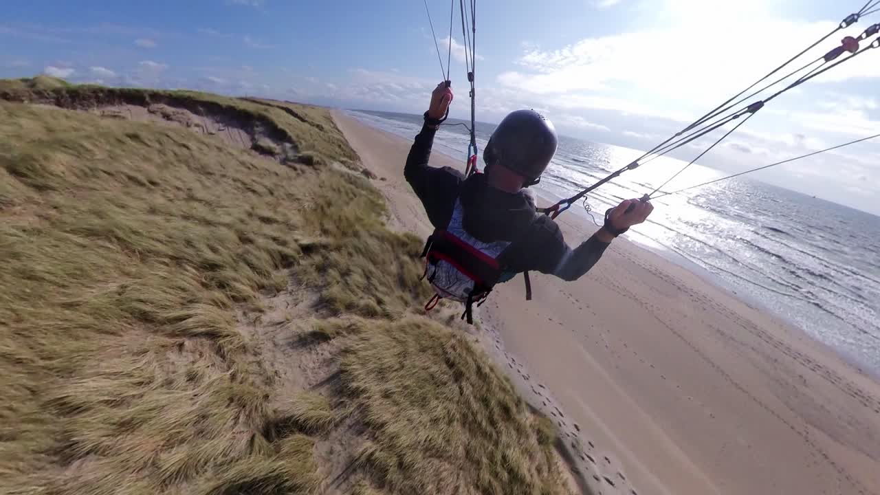Primer plano de un parapentista rozando la hierba de la duna mientras realiza acrobacias extremas, asombrosas y atrevidas.