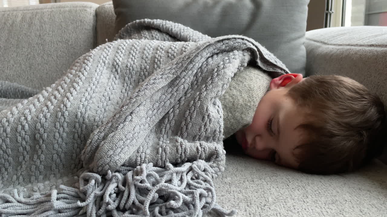 Close up of cute little boy sleeping under blanket on couch during daytime. Static
