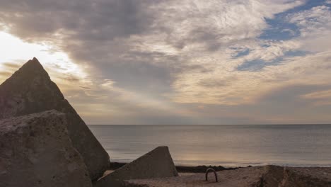 Time-lapse-of-fast-moving-clouds-over-the-Baltic-sea-at-Liepaja,-wave-breaker-in-the-foreground,-calm-sea,-sun-shining-through-the-clouds,-lightrays-shining,-copy-space,-low-angle-wide-shot