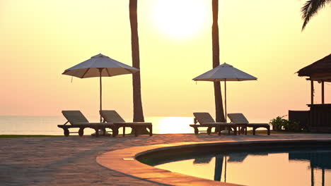 sun loungers and beach umbrellas sit empty along the edge of a resort swimming pool while the sun sets on the ocean horizon