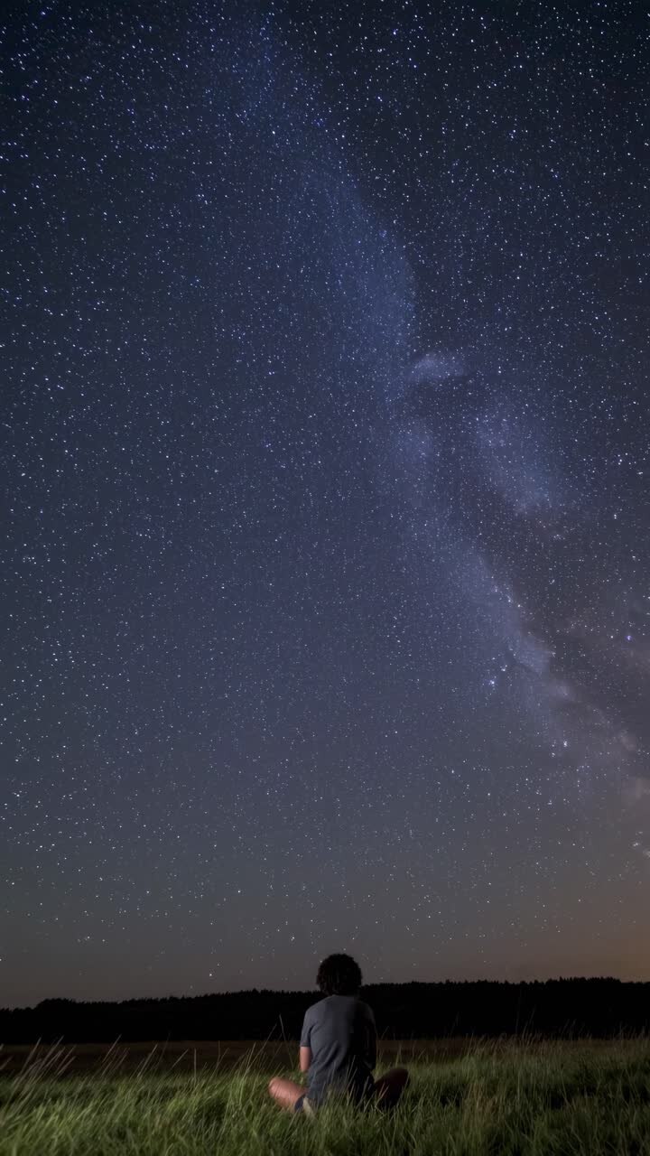 A person sits in a field gazing at a star-filled night sky. The low-angle shot captures the vastness
