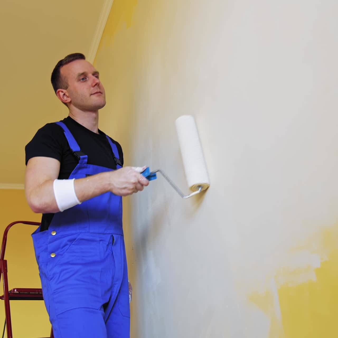 Painter man painting wall with a roller. Portrait of a young worker in overalls doing makeover with new color in the room. Professional painting services