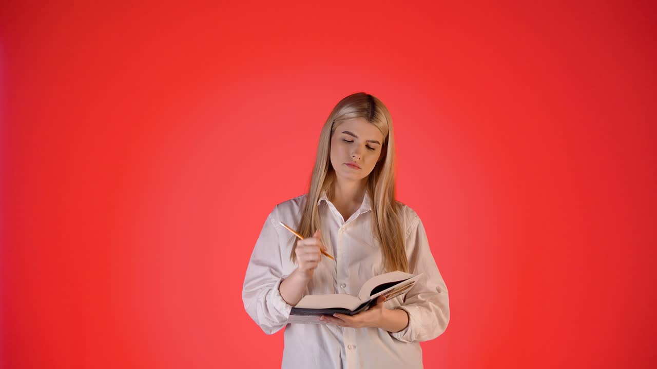 joven rubia pensativa tomando notas en un cuaderno de papel, fotografía de estudio con fondo rojo