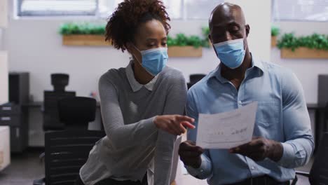 diverse male and female business colleagues in face masks talking, man holding document in office