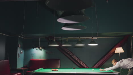 billiards player in white shirt leans over green pool table, focusing intently on his shot at the cue ball. dimly lit game room with dark walls creates a relaxed, casual atmosphere