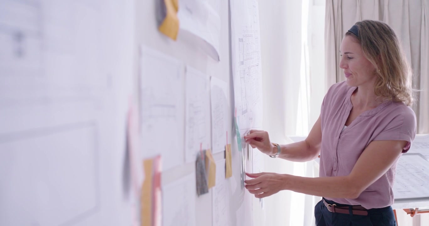 Mulher de meia-idade após revisão do conselho, vestindo blusa rosa em estúdio, movendo post-its, com espaço para texto.