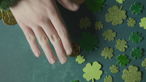 Overhead-Studioaufnahme-Einer-Hand,-Die-Goldmünzen-Neben-Kleeblattformen-Im-Hintergrund-Sammelt,-Um-Den-St.-Patricks-Day-Zu-Feiern