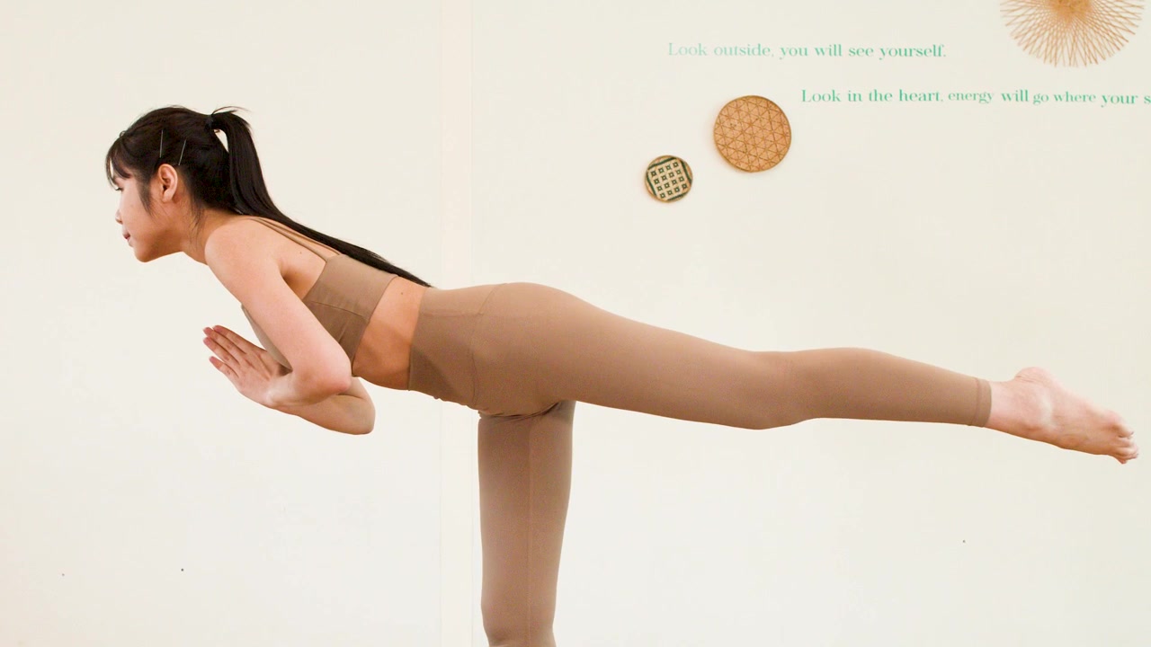 An Asian woman practices the warrior three yoga pose in a bright minimalist studio setting