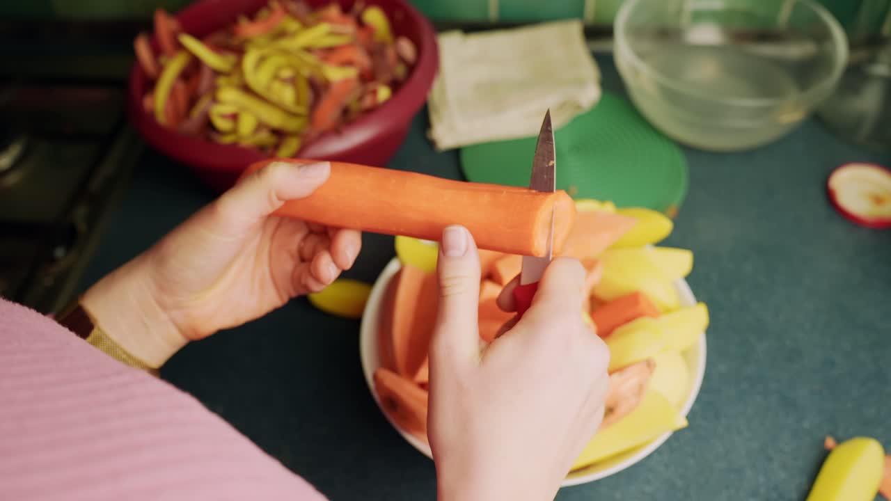 Mani in primo piano affettano una carota con un piccolo coltello sopra una ciotola di verdure pretagliate su un bancone da cucina, mostrando una semplice preparazione casalinga