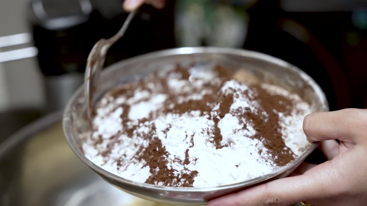 Close up of a pastry chef's hands mixing flour and cocoa powder with a spoon in a metal bowl, preparing ingredients for baking a chocolate cake