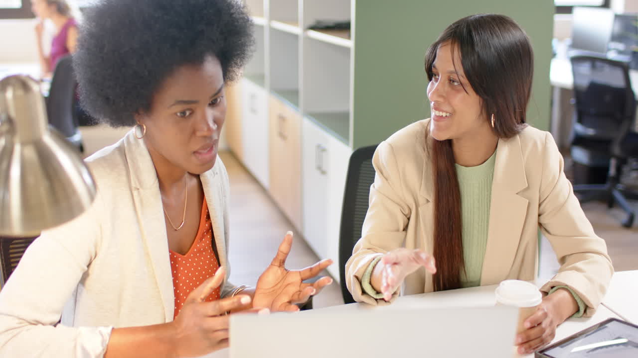 Happy diverse female creative colleagues in discussion using laptop in office, slow motion