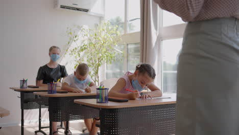 At-school-in-the-classroom-children-write-in-notebooks-in-protective-masks-observing-a-social-distance.-School-education-during-the-pandemic