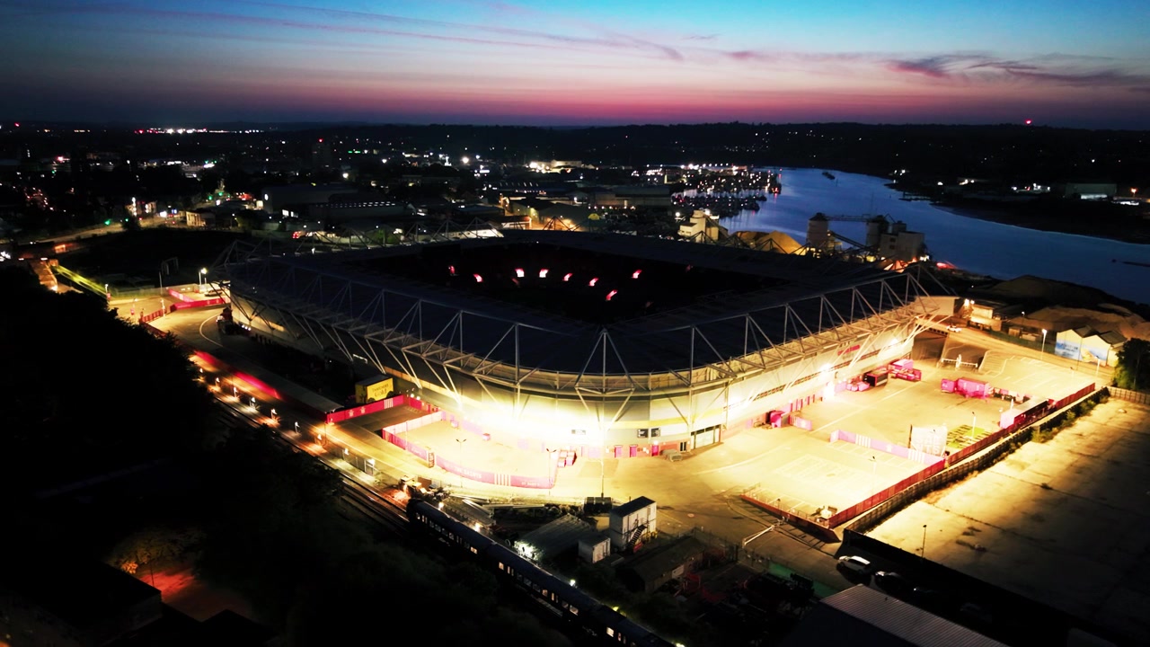 Close drone view of brightly illuminated St Marys Stadium as city lights fade beneath a blue and pink early morning sky