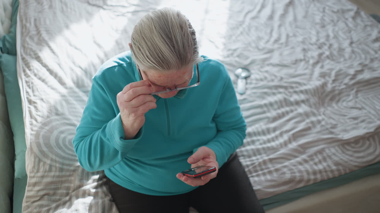 Elderly woman in turquoise fleece sweater sitting on bed, focused on her mobile phone, holding it with one hand, with her head slightly bent as she looks down. Soft natural light in background