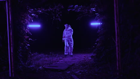 person in white suit in a dark forest at night with purple lights
