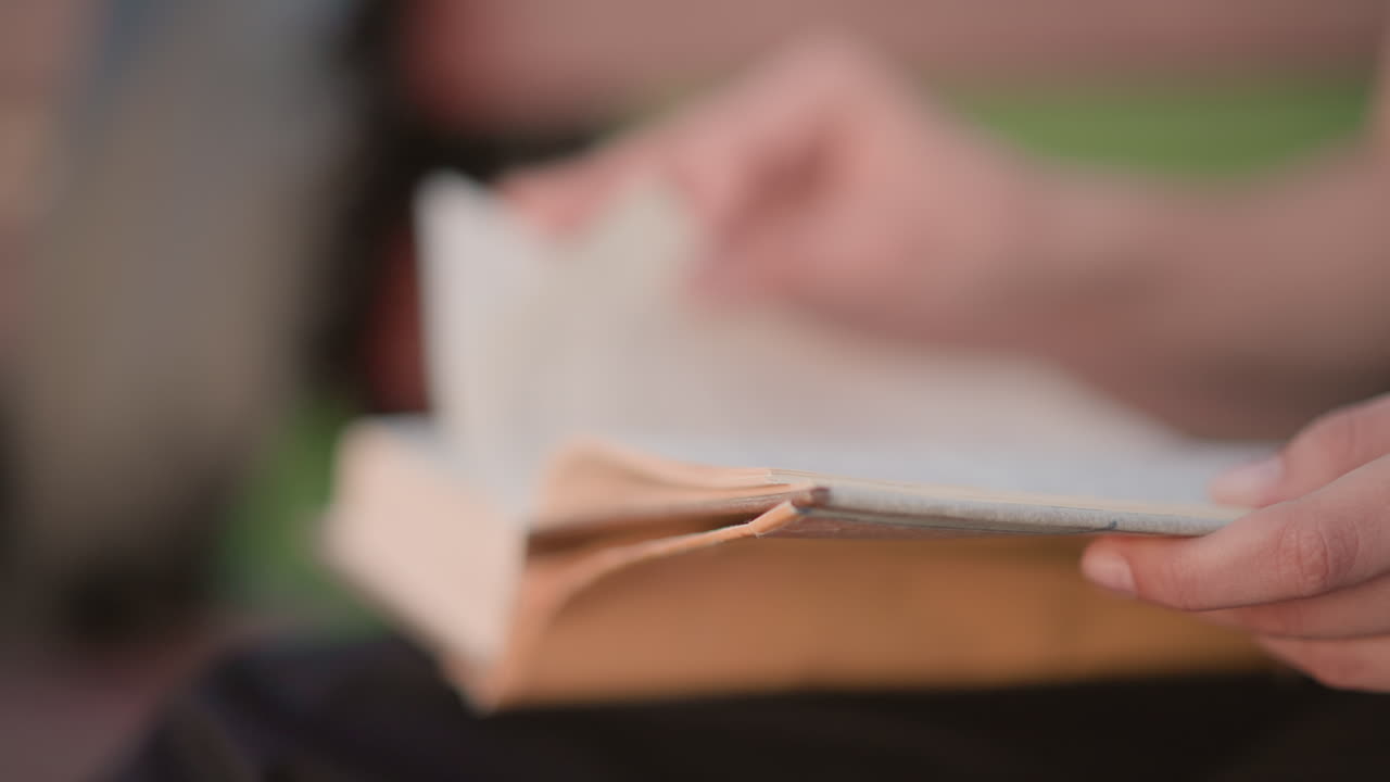 Hænder der bladrer på en bænk, aften i byen i baggrunden, tommelfingeren markerer linjen, slidt paperback hviler i skødet, bypendler holder en stille pause mellem transport, blød neonglans, lav dybdeskarphed, diskret